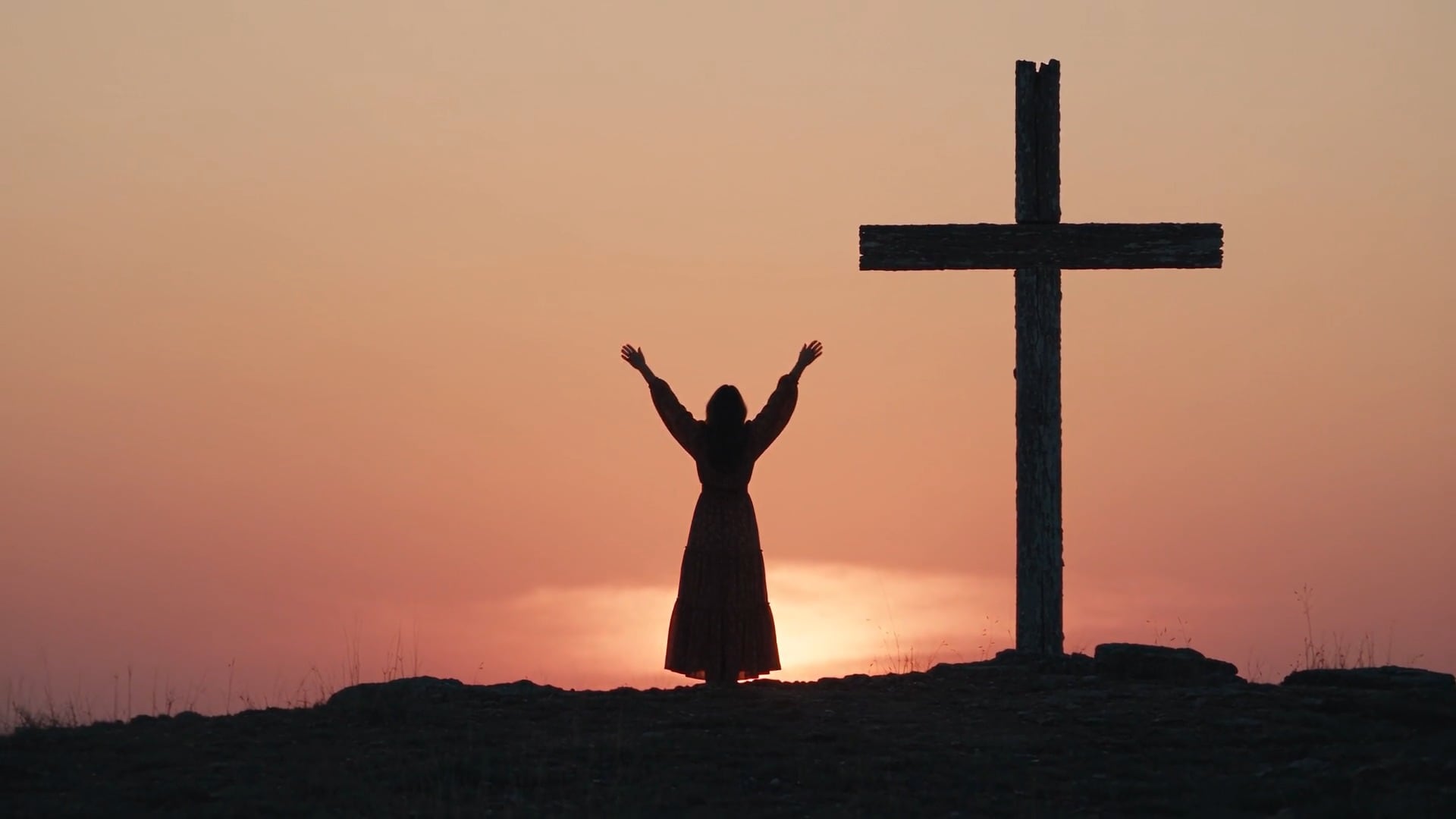 A woman with her arms raised in praise beside a wooden cross at sunset